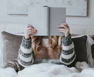 Frau sitzt im Bett mit einem Buch vor dem Gesicht